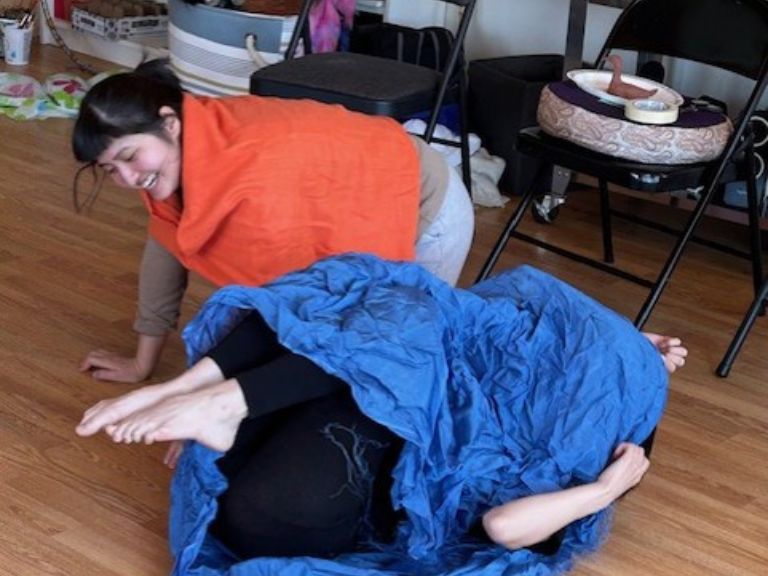 Two people on floor, one person on hands and knees and one person on back wrapped in blue material.