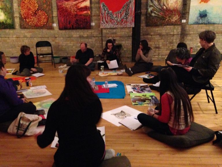 Group of people sitting in circle on chairs and on the floor working on art projects.