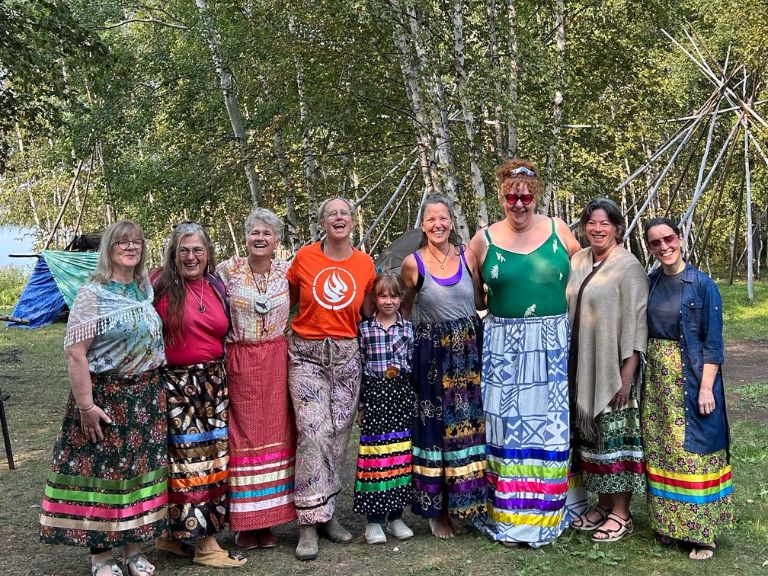 Group of people standing in front of tipi structures and forest.